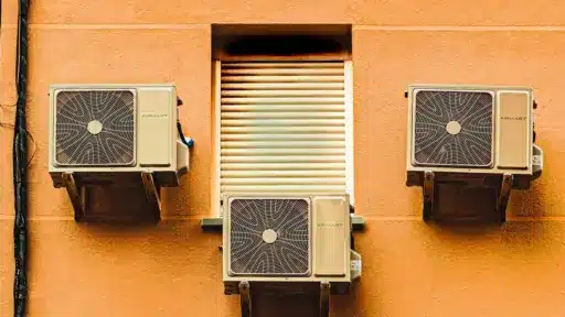 Three air conditioning units mounted on an orange exterior wall with a closed shuttered window