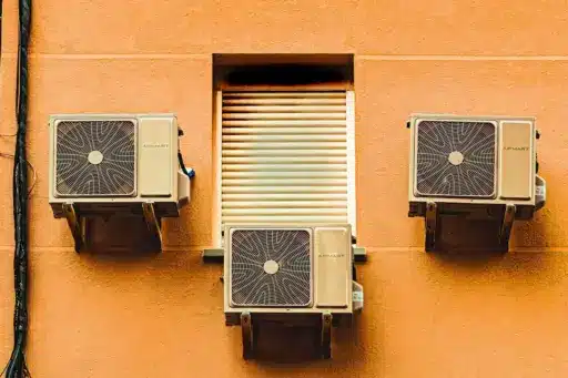 Three air conditioning units mounted on an orange exterior wall with a closed shuttered window