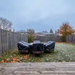Outdoor patio furniture with protective covers on frosty lawn surrounded by wooden fence in fall setting
