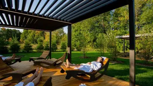 Person relaxing on wooden lounge chair under pergola in sunny garden setting