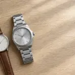 Two wristwatches with leather and metal bands on a light wooden surface under soft sunlight