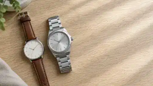 Two wristwatches with leather and metal bands on a light wooden surface under soft sunlight