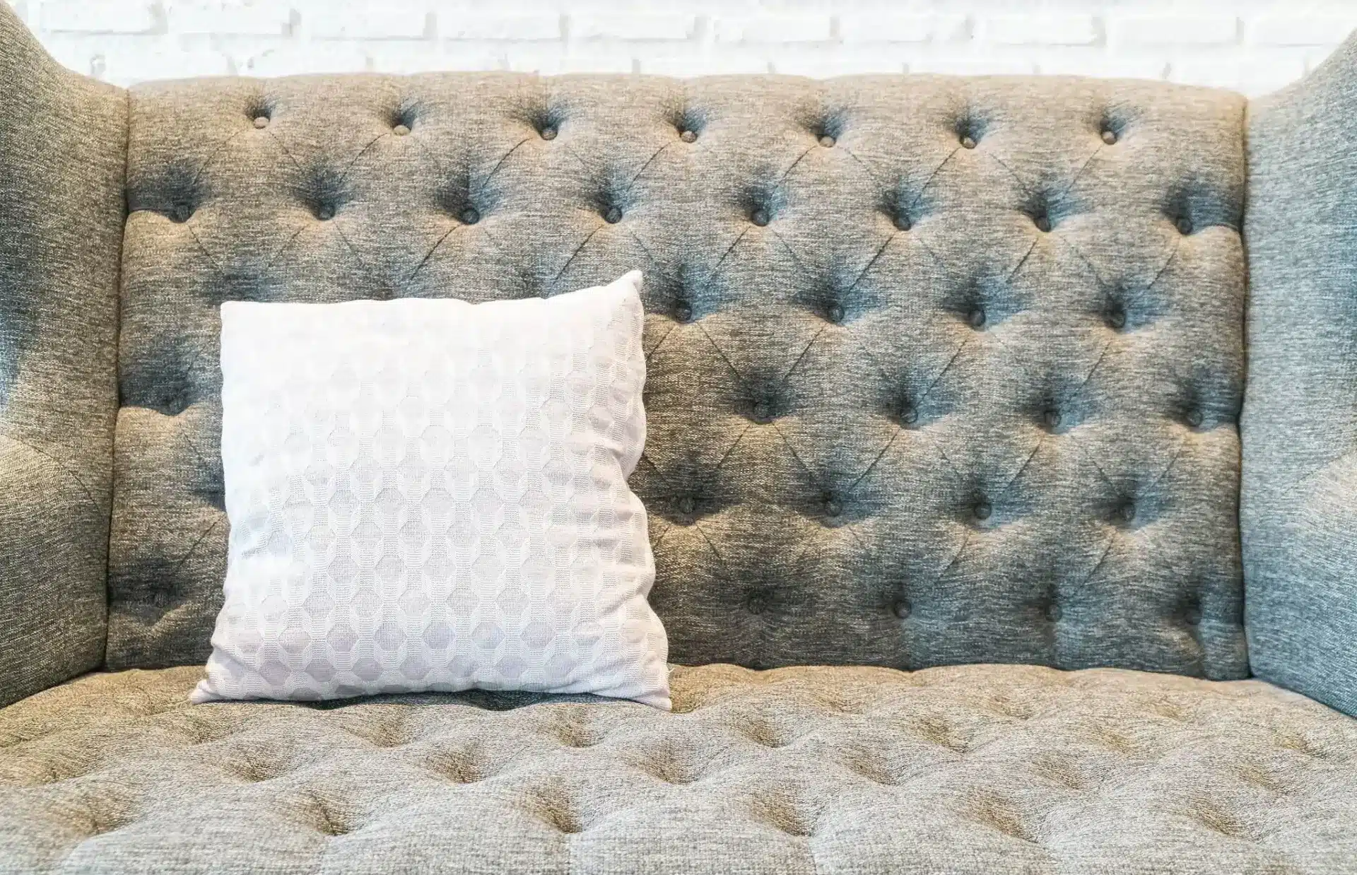 Gray tufted sofa with white patterned pillow against white brick wall backdrop