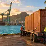 Dock workers securing cargo container on truck at sunset near harbor cranes and ocean backdrop