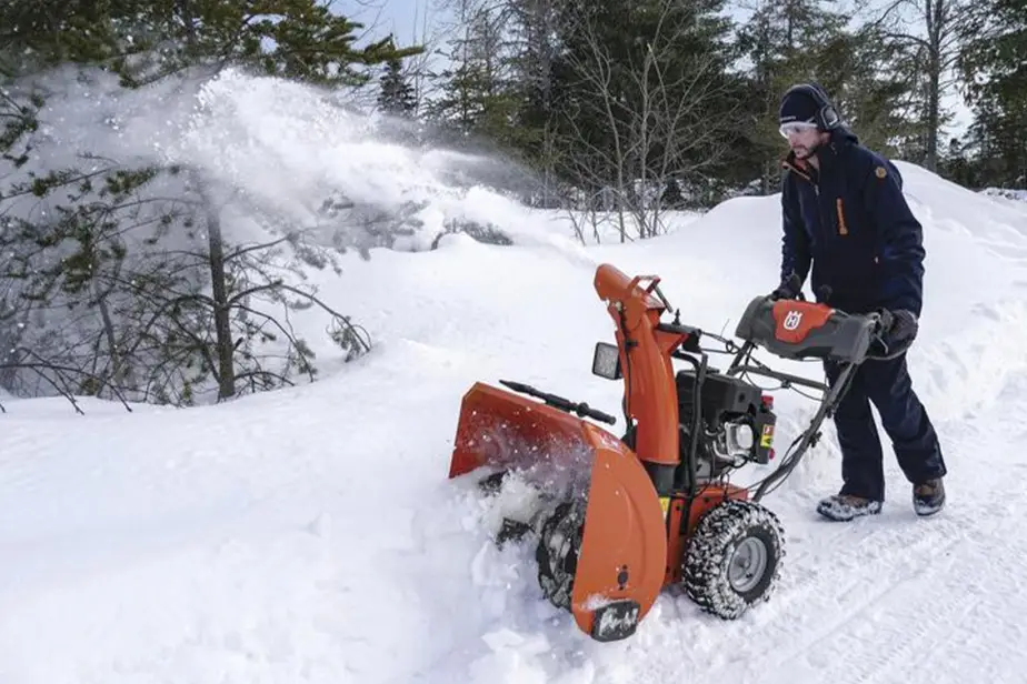 Person using an orange snow blower to clear snow on a snowy path in wooded area