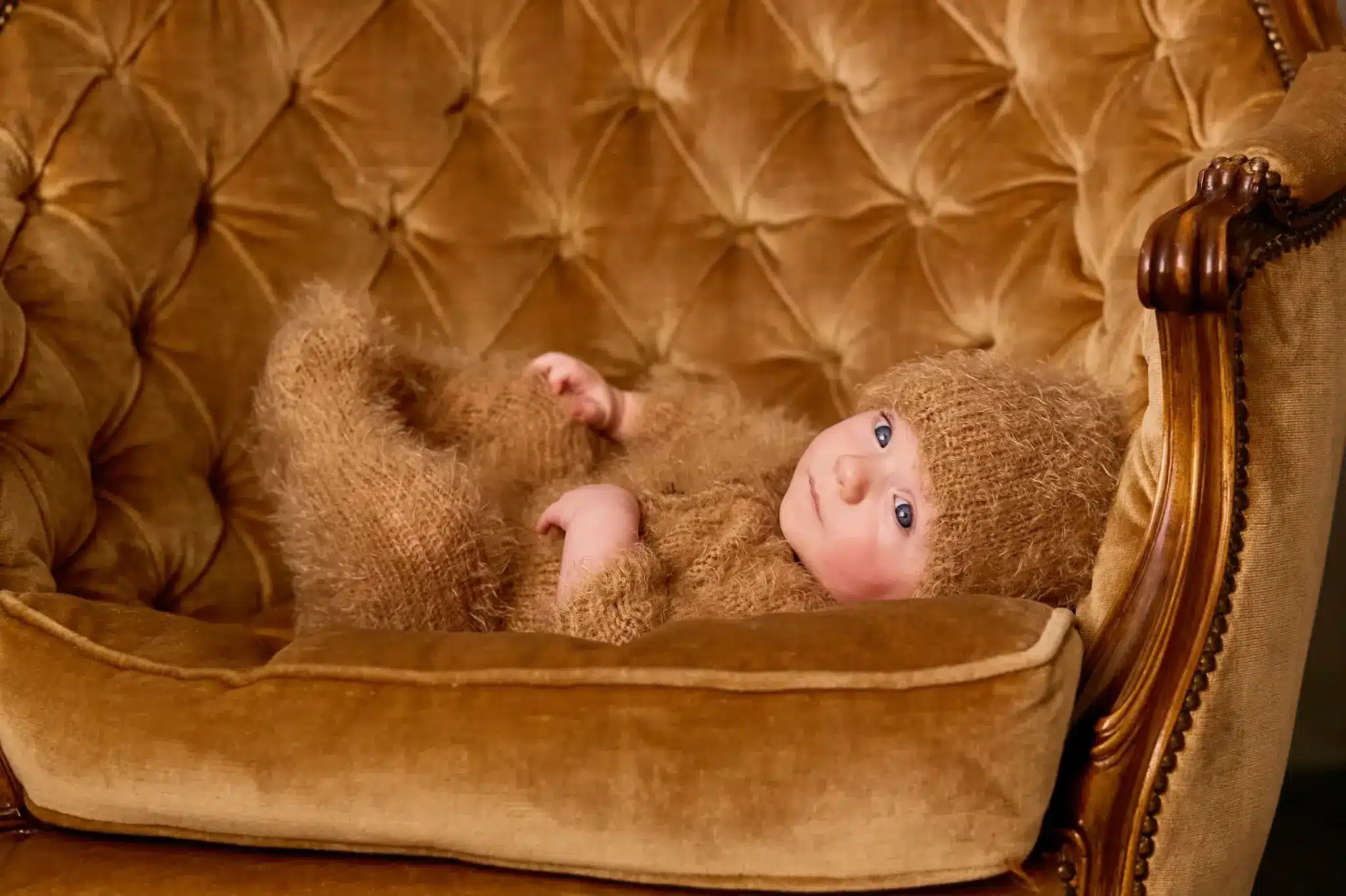 Baby in fuzzy brown outfit lying on vintage tufted armchair indoors
